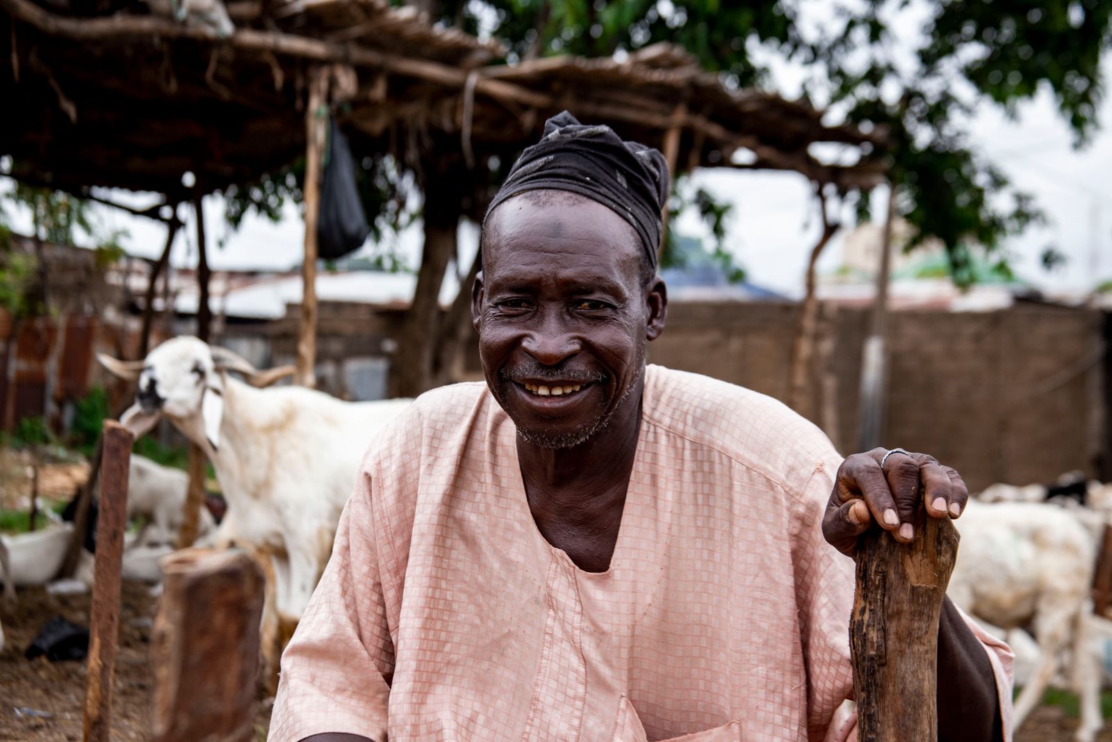 A farmer smiling in front of his goats, under a thatched shelter
